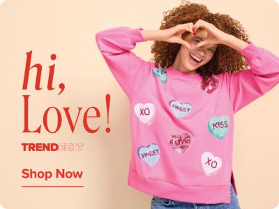 A woman wearing a pink candy heart sweatshirt. Hi, love! Trendedit. Shop now.