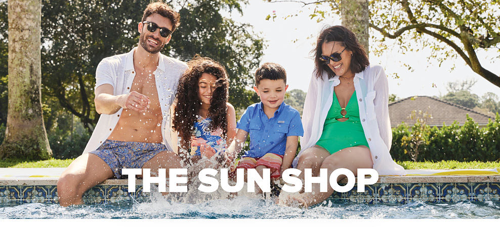 A family in swimwear sits on the edge of a pool, splashing their feet in the water. The Sun Shop.