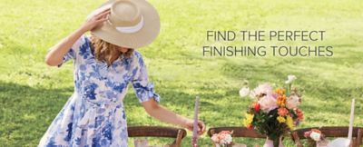 A woman in a blue and white floral dress holds her straw hat while setting a table outside. Find the perfect finishing touches.