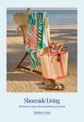 A beach chair with a towel and sunhat placed on top next to a tote bag and sandals. Shoreside Llving. Sit back & relax with everything you need. Shop outdoor living.