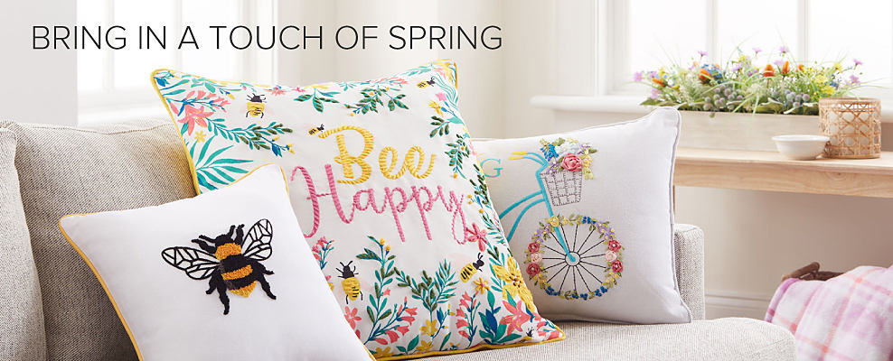 An image of a couch with spring throw pillows on it, including one that says Bee Happy. Bring in a touch of spring.
