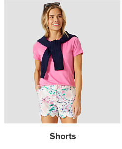 A woman in a hot pink tee, patterned shorts with a navy crewneck tied around her neck. Shorts.