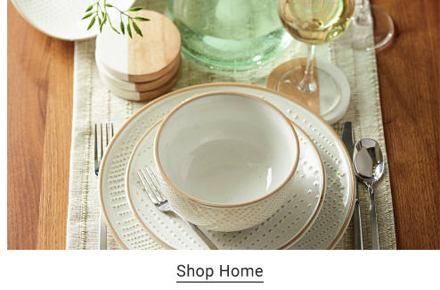 On a table sits white dinnerware with brown rims, including a plate, salad plate and bowl, flanked by cutlery and with wood coasters and a wine glass. Shop Home. 