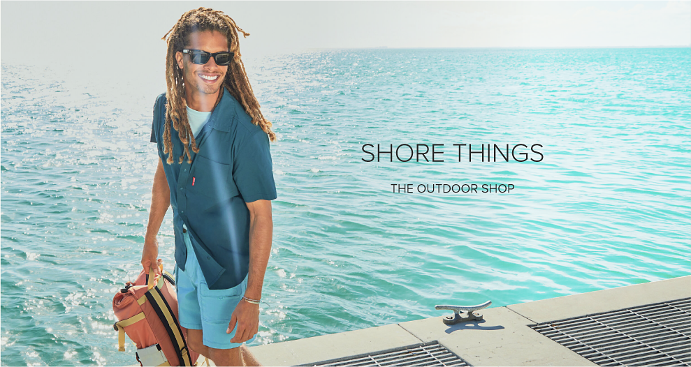 A man stands on a concrete dock. He wears a blue short sleeve button up shirt, with a lighter blue pair of shorts. He holds an orange Topo Designs backpack in his hand. Shore Things. The Outdoor Shop. 