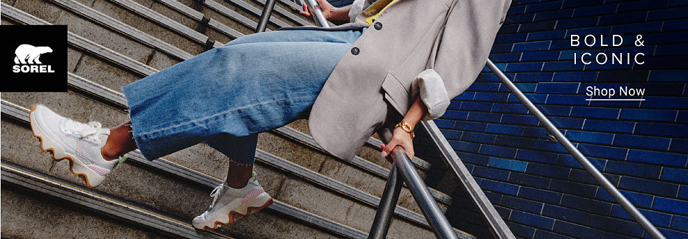 Sorel logo. Image of a woman in a gray coat, jeans and white sneakers. Bold and iconic. Shop now.