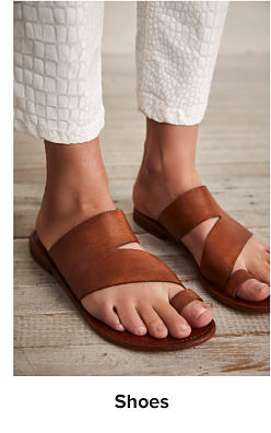 A woman wearing white ankle length pants with brown slip on sandals. Shop shoes.