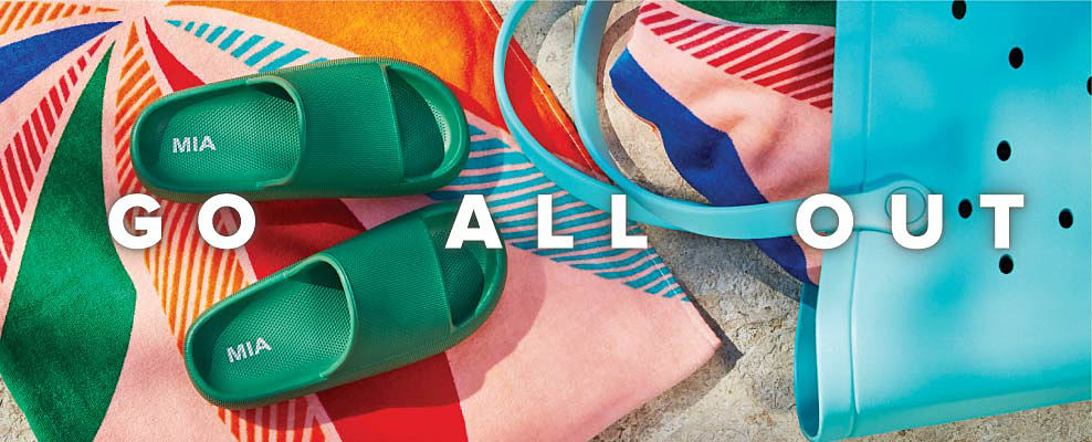 An image of a pair of green slip on sandals sitting on a bright beach towel, beside a blue beach bag. Go all out. 