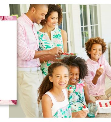 A man wearing a pink & white plaid long sleeved button front shirt & beige pants standing next to a woman wearing a multi colored tropical print sleeveless dress, a girl wearing a multi colored tropical print sleeveless dress, a girl wearing a multi colored tropical print sleeveless dress & a boy wearing a pink & white plaid long sleeved button front shirt & beige pants.