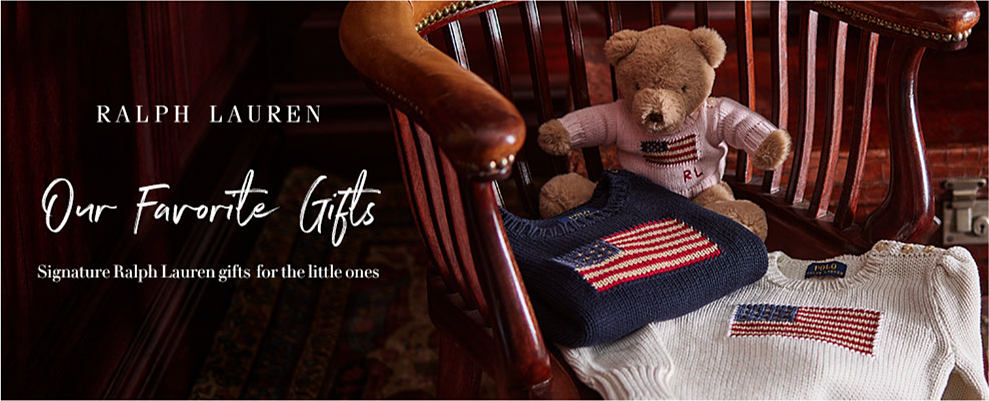 A small teddy bear sits in a chair, with folded blue and white sweaters with the American flag on them. Ralph Lauren. Our favorite gifts. Signature Ralph Lauren gifts for the little ones. 