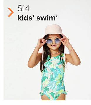 A girl in a one piece swimsuit with green palm tree leaves on it. She also wears sunglasses and a pink bucket hat. $14 kids' swim. 