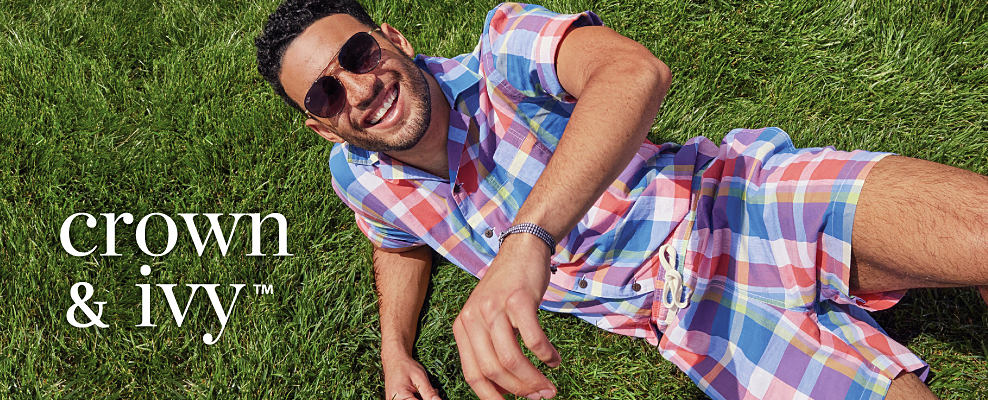 A man laying in the grass wearing a red, white and blue plaid short sleeve button front shirt and matching short and sunglasses. The Crown and Ivy logo.