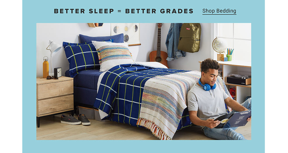 BETTER SLEEP equals BETTER GRADES, Shop Bedding. A young man sitting on the floor in his dorm room, wearing headphones around his neck and holding a vinyl album. His bed is blue with a green windowpane print. There's a side table and a variety of decor. Shop Bedding. 