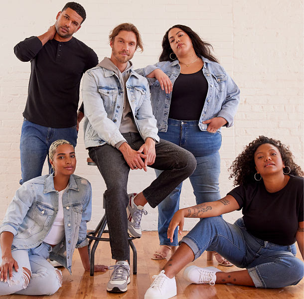 Two men and three women pose in front of a white brick wall. One man wears a black henley shirt and blue jeans. One man wears a light wash denim jacket over a gray hoodie and black jeans. One woman wears a light wash denim jacket over a black shirt and blue jeans. One woman wears blue jeans, white sneakers and a black t-shirt. One woman wears distressed light-wash jeans and a light wash denim jacket over a white t-shirt. 