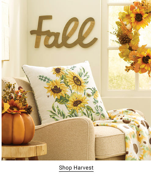 An image of a sunflower throw pillow on a chair, with a golden decor piece showing the word fall above it. Shop Harvest.