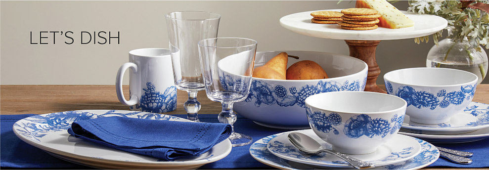 A wooden table topped with white dinnerware with blue floral designs. Two stemmed wine glasses sit beside a bowl of pears and a small tray of crackers and cheese. Let's dish. 