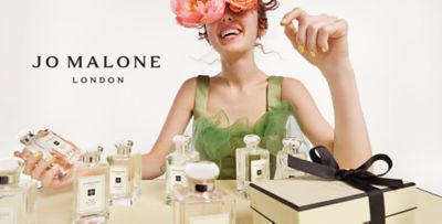 An image of a woman wearing a green top sitting at a table topped with Jo Malone fragrances. The Jo Malone London logo.