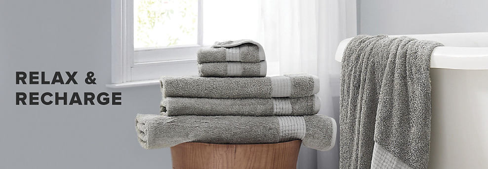 An image of a stack of folded gray hand, wash and bath towels beside a white tub. Relax and recharge.