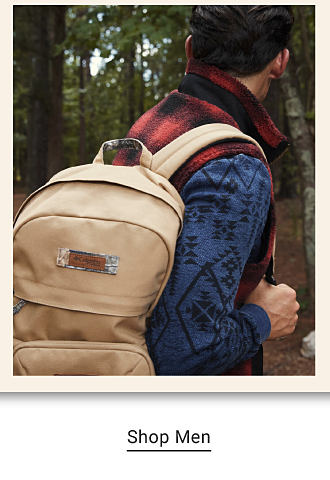 A man wearing a blue and black tribal print long sleeve top and carrying a beige backpack. Shop men.