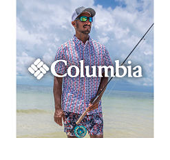 A man in a plaid Columbia button up short sleeve shirt and shorts stands on at the beach with a fishing rod. Columbia.