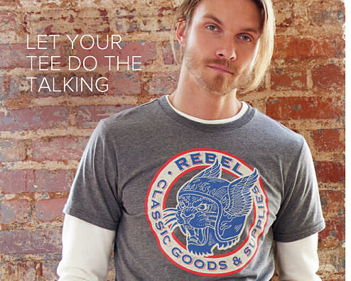 An image of a man standing in front of a brick wall, wearing a gray tee with a design of a cat wearing a winged motorcycle helmet in red, white and blue. It reads rebel classic goods and supplies. 