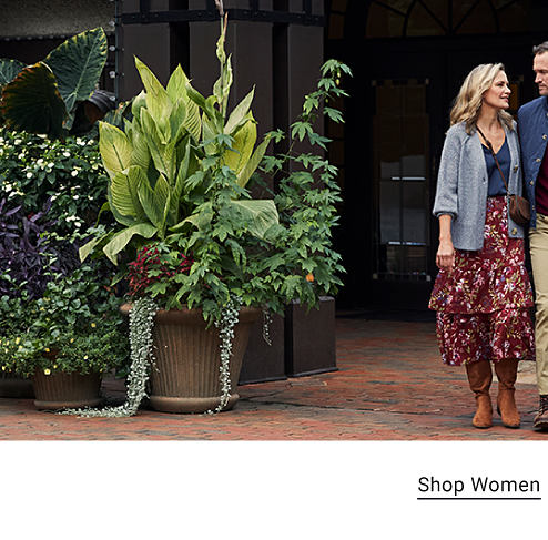 A woman wears a red floral skirt, blue top and a light blue cardigan. She walks with a man who is rolling a briefcase and wearing a red long sleeve shirt, blue vest and khaki pants. They stand in front of four wine barrels. Shop women. Shop men. 