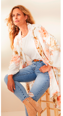 A woman in a light pink, tan and white floral duster cardigan with a white tee underneath, light jeans and tan booties.