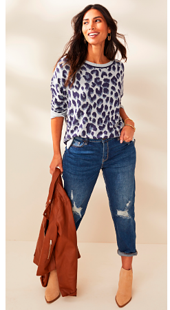 A woman in a navy blue and white leopard print sweater with ripped jeans and tan booties holding a camel colored leather jacket.