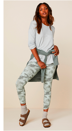 A woman in a plain off white long sleeve tee with a sage tee tied around her waist and light green camo leggings.