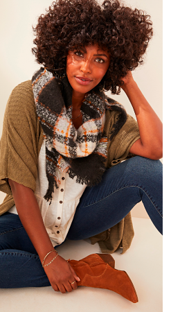 A woman in a black, orange and white plaid scarf with a white button up peplum shirt and olive short sleeve sweater with jeans and camel booties.
