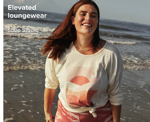 A woman standing in front of the ocean wearing a white tee with a pink and orange sunset graphic and light pink camo leggings. Elevated loungewear. Shop studio.