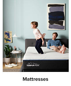 A little girl jumping on a bed with her dad sitting on the bed in the background. Shop mattresses.