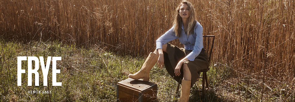 Frye. Since 1863. Image of a woman in a button down top, skirt, and boots sitting in the middle of a wheat field.