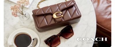 A brown leather Coach handbag, sitting beside brown sunglasses and a cup of coffee. Coach New York.