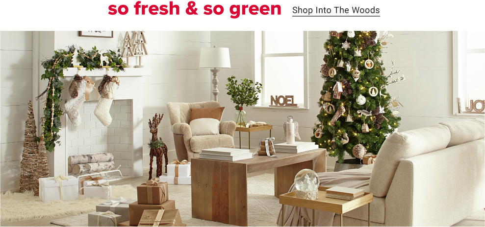 An all white room simply decorated for christmas. A decorated christmas tree sits between two windows that have holiday signs in them, one reading noel. A wooden reindeer sits beside the fire that is decked with two white stockings and wooden outlined christmas trees and greenery on its ledge. So fresh and so green. Shop into the woods.