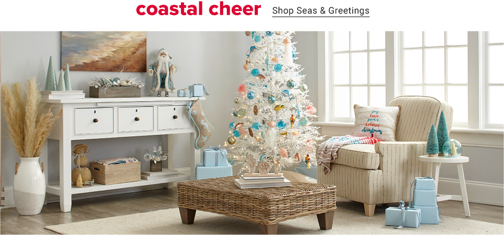 A white beach themed room has a collection of blue and pink chrismtas decorations. The white tree has an assortment of pink and blue ornaments. A cream love seat has a striped tree decorative pillow. Small green christmas trees stand on a side table. Coastal cheer, shop seas and greetings. 