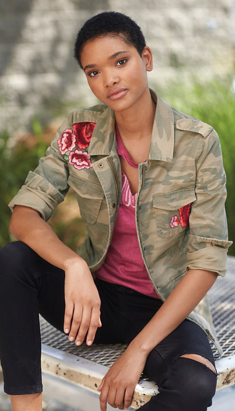 A young woman wearing a distressed red graphic tee, distressed black jeans & a green camo jacket with rose applique detail. Fall jackets. Shop now. .