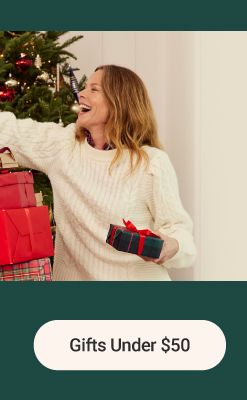 Image of a man and woman holding presents in front of a Christmas tree. The gift shop. Gifts under $50.