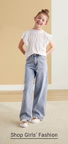 A girl in a white ruffled top, baggy blue jeans and fashion sneakers. Shop girls' fashion.