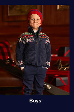 An image of a young boy in a Fair Isle-printed jacket, navy blue pants, and a red hat. Shop boy's clothing.