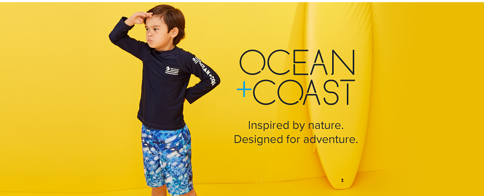 A little boy in a navy long sleeve Ocean and Coast tee shirt and blue patterned swim trunks. Ocean and Coast. Inspired by nature. Designed for adventure 