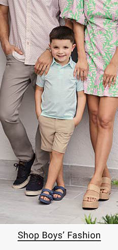 A boy in a light blue polo, khaki shorts and sandals stands between his parents. Shop boys' fashion.